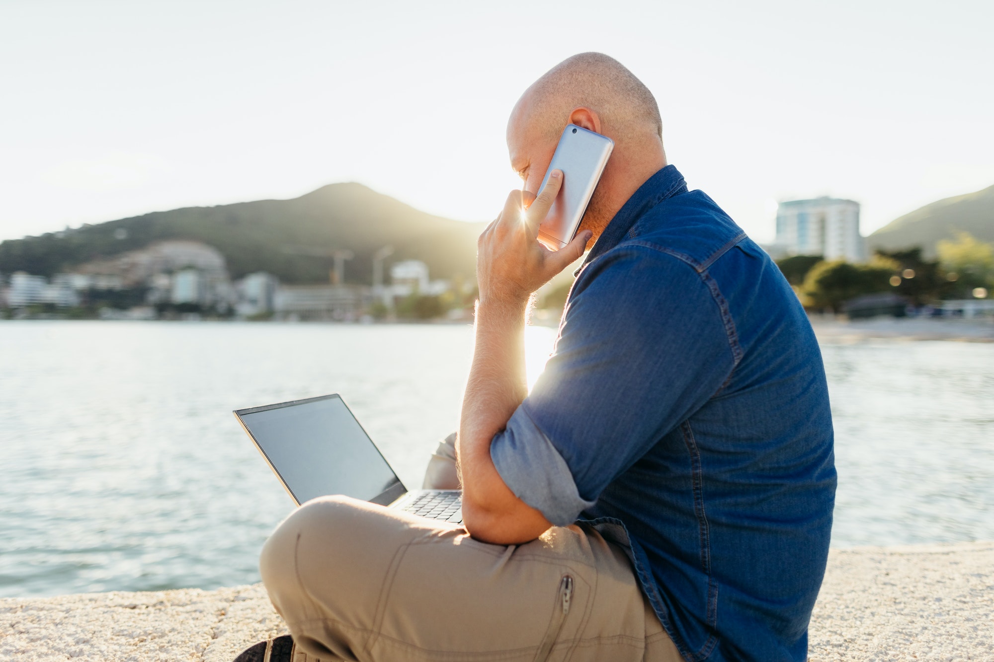 Self employed man using phone and laptop consulting clients sitting at seaside
